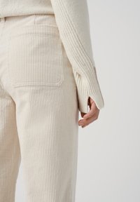 Pantalones de pana color crema con textura vertical y detalles en los bolsillos traseros, combinados con un suéter de canalé color crema claro.