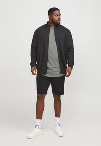 Black zip-up jacket, olive green t-shirt, black shorts, white sneakers. Socks with light blue patterns. Casual, relaxed fit.
