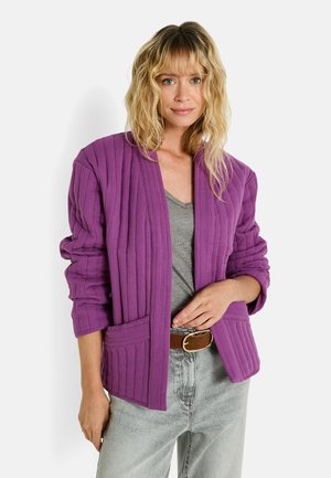 Veste violette côtelée avec une coupe décontractée, devant ouvert et deux poches avant. Portée sur un haut gris et associée à un jean clair.