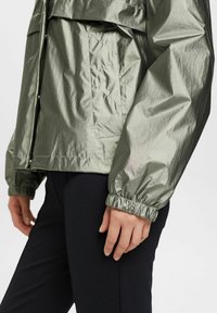 Lekka, błyszcząca oliwkowo-zielona kurtka z elastycznymi mankietami i przednią kieszenią. Gładka tekstura z lekko pomarszczonym wykończeniem.