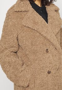 Donna che indossa un cappotto teddy color cammello con trama, ampi revers, bottoni doppiopetto e mani nelle tasche laterali sopra una maglietta nera.