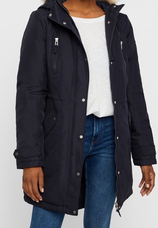 Kvinna i en svart vinterjacka med huva, dragkedja och tryckknappar, över en vit skjorta och blå jeans.