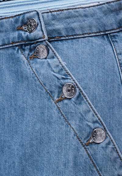 Κοντινή λήψη μπλε τζιν ύφασματος με έξι εκτεθειμένα ασημένια κουμπιά διατεταγμένα διαγώνια, κάθε κουμπί χαραγμένο με "Claudie Pierlot."