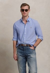 Lys blåstripet skjorte med knapper, denim jeans, brunt belte og mørke solbriller. Kjennetegn inkluderer et brystlogo og oppbrettede ermer.