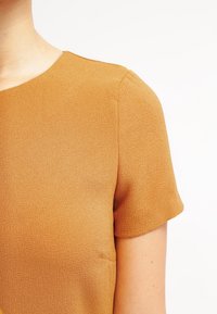 Kortärmad topp i senapsgul färg, tillverkad av textil med struktur och rund halsringning. Sömnadsdetaljer synliga på axeln och sidan.