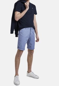 T-shirt blu navy abbinato a pantaloni corti azzurri arrotolati, scarpe da ginnastica bianche e una felpa nera appoggiata sul braccio. Texture di cotone liscia.