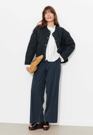 Femme portant une veste matelassée noire, un chemisier blanc, un pantalon large bleu marine à fines rayures, des sandales plateformes noires, tenant une pochette beige.