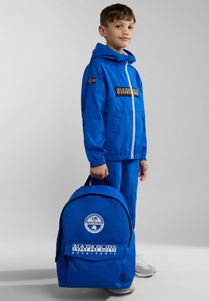 Veste coupe-vent bleue avec une fermeture éclair blanche, assortie à un pantalon. Le sac à dos bleu présente des accents de logo blancs et une forme arrondie.
