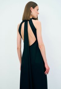 Černé šaty bez rukávů s nízkým otevřeným zadním dílem a zavazováním na krku, vyznačující se splývavou tkaninou, která elegantně padá dolů po zádech.