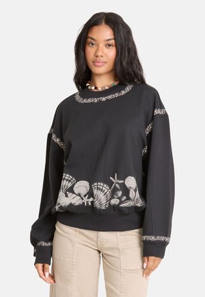 Frau mit langen dunklen Haaren, die einen schwarzen Pullover mit weißen Muschel- und Seesternmotiven und beigen Hosen vor einem schlichten Hintergrund trägt.