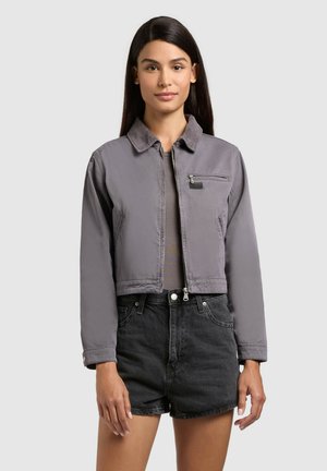 Kvinde iført en grå cropped jakke over en grå top og højtaljede sorte denimshorts, stående foran en ensfarvet lys baggrund.