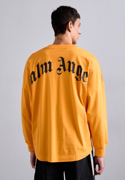 Langærmet, løs pasform t-shirt i lys orange med en rund kant. Har fed sort tekst med teksten "palm Ange" på ryggen. Lavet af bomulds-materiale.