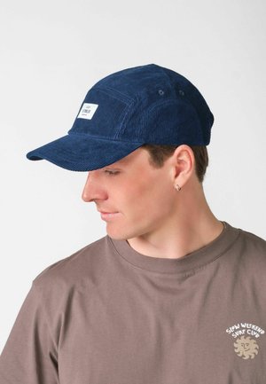 Mladý muž s modrou manšestrovou čiapkou a hnedým tričkom s logom „Slow Weekend Surf Club“, pozerajúci sa dole na svoju ľavú stranu.