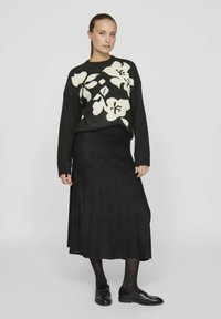 Chlupatý černý svetr s bílým květinovým vzorem, doplněný dlouhou černou sukní. Modelka má na sobě černé punčocháče a mokasíny. Jednoduchý, strukturovaný outfit.