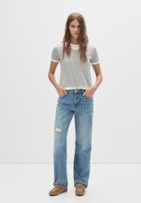T-shirt grigia a maniche corte con colletto bianco, abbinata a jeans chiari strappati. Il modello indossa mocassini color sabbia, in piedi su uno sfondo bianco.