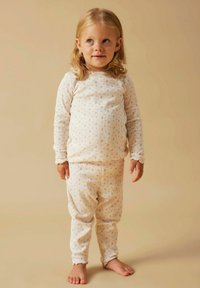 Małe dziecko z blond włosami stojące boso, ubrane w dopasowane kremowe piżamy z prążków w małe kwiatowe wzory, na beżowym tle.