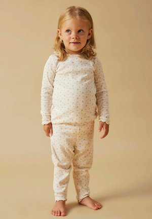 Małe dziecko z blond włosami stojące boso, ubrane w dopasowane kremowe piżamy z prążków w małe kwiatowe wzory, na beżowym tle.