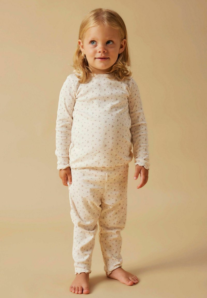 Małe dziecko z blond włosami stojące boso, ubrane w dopasowane kremowe piżamy z prążków w małe kwiatowe wzory, na beżowym tle.