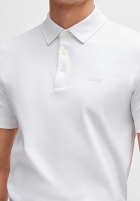 Polo blanc en tissu lisse avec une patte de boutonnage, un col plié et un logo discret embossé sur la poitrine.