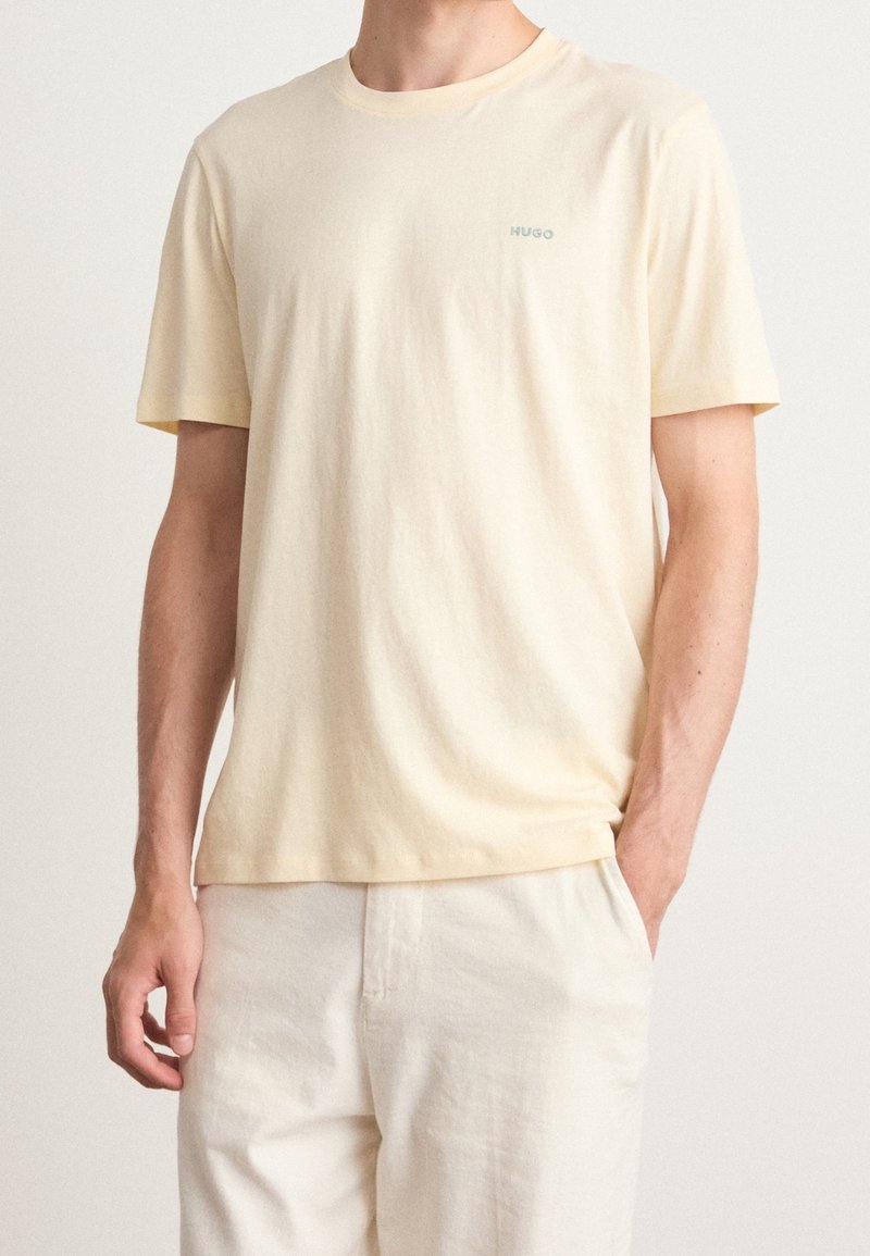 Un homme portant un t-shirt HUGO à manches courtes beige clair et un pantalon crème, avec une main dans la poche, devant un fond uni.