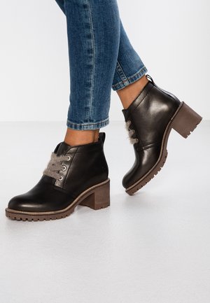 Bottines noires en cuir avec talons marron épais et lacets gris duveteux portées avec un jean bleu retroussé sur un sol blanc.