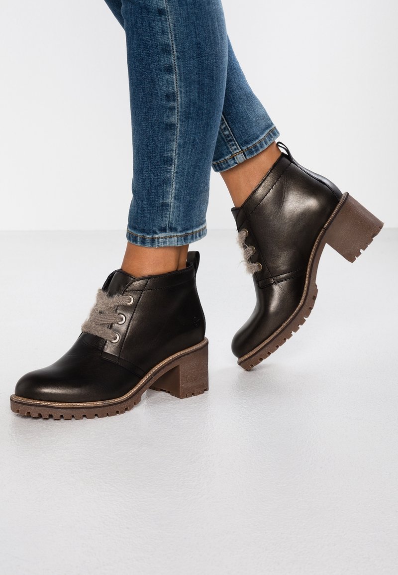 Bottines noires en cuir avec talons marron épais et lacets gris duveteux portées avec un jean bleu retroussé sur un sol blanc.