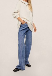 Beige übergroßer Kapuzenpullover mit geripptem Textur, kombiniert mit blauen weiten Jeans. Das Model trägt schwarze Slipper, die Hände in den Taschen.