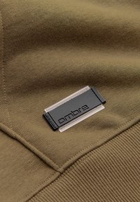 Nahaufnahme von olivgrünem Stoff mit einem schwarz-weißen rechteckigen „Ombre“-Markenlabel, das nahe einer Naht aufgenäht ist.