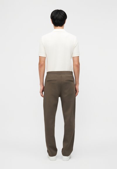 Άνδρας που στέκεται στραμμένος προς τα πίσω, φοράει λευκό πουκάμισο με κοντά μανίκια, καφέ παντελόνια με πίσω τσέπες και λευκά παπούτσια σε απλό φόντο.