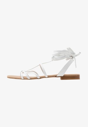 Sandale plate en cuir blanc avec des lanières fines sur les orteils, un lien à nouer autour de la cheville et un petit talon beige.