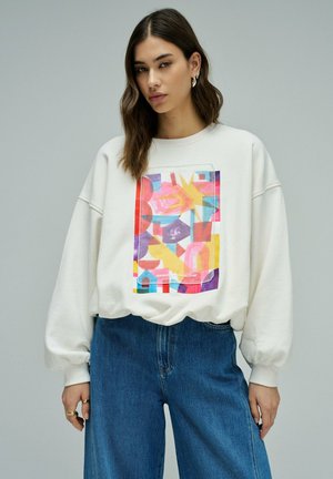 Femme portant un sweat-shirt blanc oversize avec un motif rectangulaire abstrait coloré et un jean bleu, debout devant un fond gris uni.