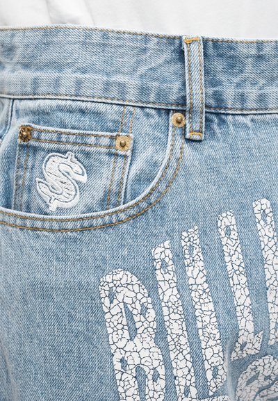 Τζιν παντελόνι από ανοιχτό μπλε τζιν με ξεθωριασμένο φινίρισμα, που διαθέτει ένα εμφανές σπασμένο λευκό κείμενο και έναν κεντημένο σύμβολο δολαρίου στην τσέπη.