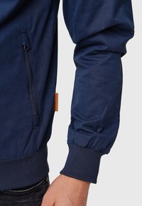 Manche de veste bleu marine avec poignet côtelé, poche latérale zippée visible et une petite étiquette de marque en cuir marron.
