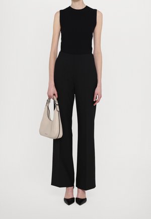 Persoon in een mouwloos zwart topje en hoge zwarte broek, met een beige handtas in de linkerhand en zwarte puntige schoenen.