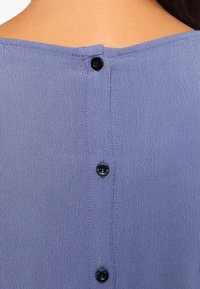 Violetti pusero, jossa on teksturoitu kangas ja napitus takana. Mustat napit ja sileä, pehmeä tekstuuri korostavat sen muotoilua.