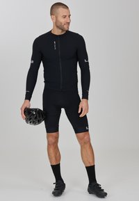 Zwart fietsoutfit met een langemouwen top met een ritssluiting aan de voorkant en zwarte shorts. Gemaakt met zwarte fietsschoenen en houdt een zwarte helm vast.
