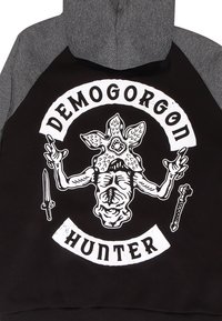 Felpa nera con maniche grigie presenta una grafica bianca di una creatura intitolata "DEMOGORGON" con spade e la parola "CACCIA" sotto.