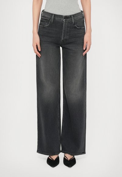 Persoon in hoge taille brede zwarte vervaagde jeans en zwarte puntige hakken met dunne banden tegen een witte achtergrond.