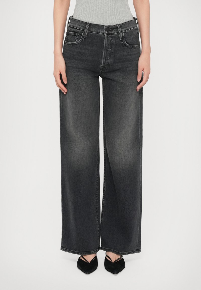 Oseba nosi hlače z visokim pasom in širokimi nogavicami v črni barvi, ki so poudarjene s črno koničasto obutvijo s tankimi trakovi, na beli podlagi.