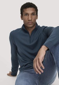 Dunkelblaues Reißverschluss-Sweatshirt mit geripptem Struktur, das einen hohen Kragen hat. Kombiniert mit blauen Denim-Jeans. Der Stoff wirkt weich und komfortabel.