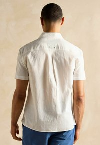 Kratkām piedurknēm, ar jostu, balts lina krekls ar smalku tekstūru; tam ir dekoratīvas pogas un aizmugurējais plisējumam, lai nodrošinātu labu piegriezumu.