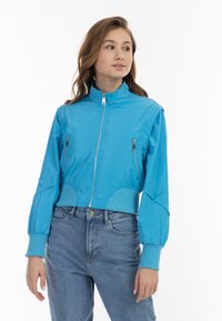 Giacca bomber azzurro chiaro, tagliata, con colletto alto, tasche con zip e polsini elasticizzati. Tessuto liscio e lucido con una vestibilità attillata.