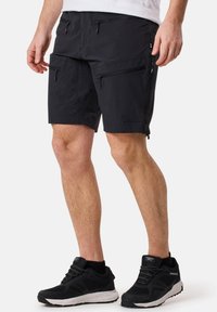 Schwarze Outdoor-Shorts aus leichtem Material mit mehreren Reißverschlusstaschen und einem bequemen Schnitt. Kombiniert mit schwarzen Sportschuhen.