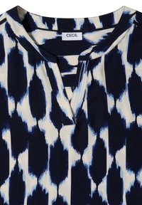 Col et encolure d'un chemisier à motifs noir et blanc avec une teinte bleutée, comportant une petite étiquette en tissu indiquant "CECIL."