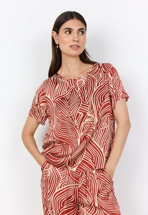 Kvinna med långt mörkt hår som bär en matchande röd och beige abstrakt mönstrad kortärmad topp och byxor, står med händerna i fickorna.