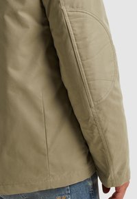 Ljusolivgrön jacka med texturerad yta, vadderade armbågsskydd och en enkel, strömlinjeformad design. Synliga sömmar längs med sömmarna.