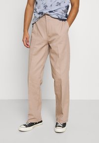 Pantalones beige claros de corte relajado, con pliegues frontales, bolsillos laterales y una textura de tela suave, combinados con zapatillas negras.