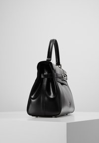 Black leather handbag with structured shape, top handle, silver hardware, and decorative chain on a white pedestal against a gray background.
