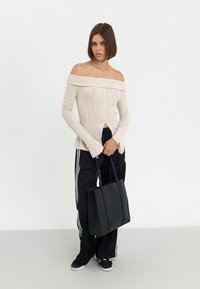 Světle béžový top bez ramínek s ribovanou texturou, doplněný černými sportovními kalhotami s bílými pruhy, držící černou tašku.