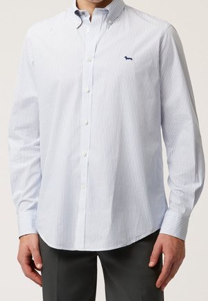 Mand iført lyseblå og hvide lodrette stribet skjorte med knapper og mørkt gravhundlogo på brystet og mørke bukser.